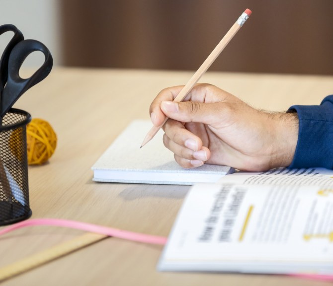 Henkilön kädessä on kynä ja pöydällä on avoin opas.