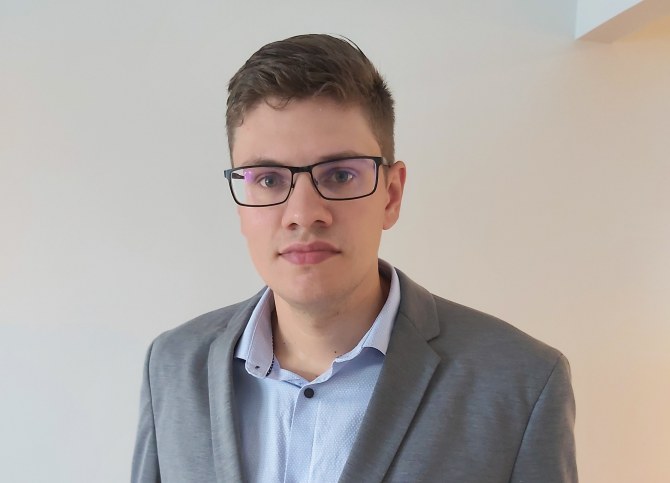 Tapio Helotie seisoo valkoisen seinän edessä ja katsoo kameraan.