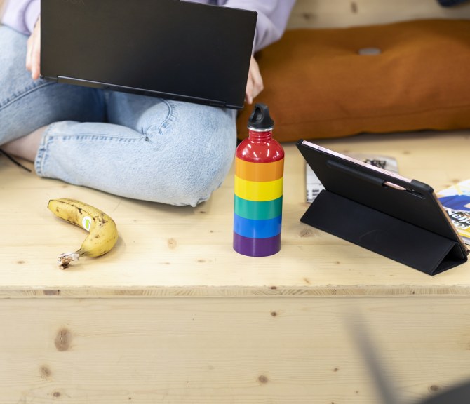 Henkilö istuu portaalla läppäri sylissään, vieressä sateenkaaren värinen juomapullo.