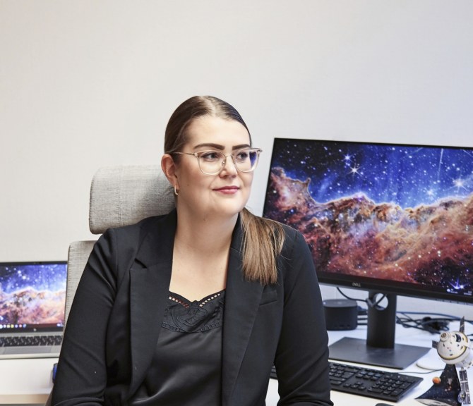 Kiira Tiensuu istuu tietokoneen edessä, tietokoneen näytöllä on avaruusmaisema.