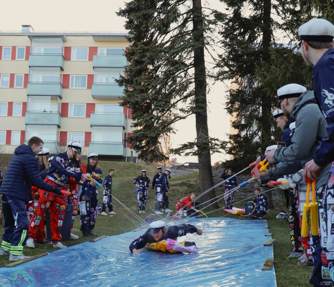 Teekkari laskee maata vasten laitetusta eristemuovista tehtyä liukumäkeä pitkin samalla kun muut ampuvat häntä kohden vesipyssyillä.