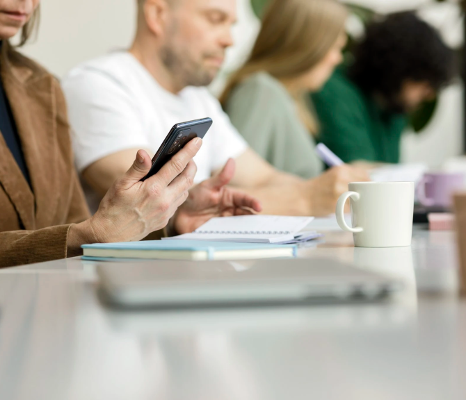 Neljä henkilöä istumassa pöydän ääressä keskittyen omiin tehtäviinsä. Etualalla olevalla on kädessään puhelin ja edessään kannettava tietokone.