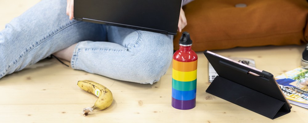 Henkilö istuu portaalla läppäri sylissään, vieressä sateenkaaren värinen juomapullo.