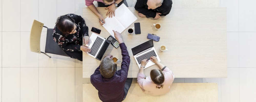 Työporukka pöydän ääressä, ylhäältä päin kuvattuna.