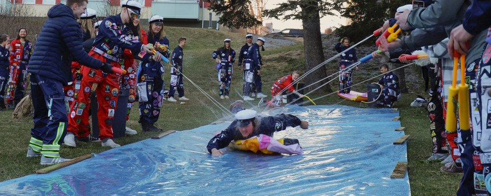 Teekkari laskee maata vasten laitetusta eristemuovista tehtyä liukumäkeä pitkin samalla kun muut ampuvat häntä kohden vesipyssyillä.