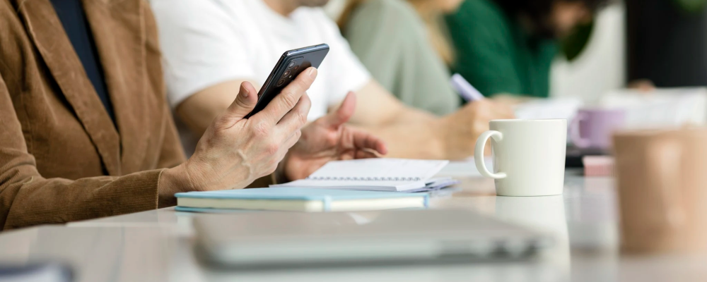 Neljä henkilöä istumassa pöydän ääressä keskittyen omiin tehtäviinsä. Etualalla olevalla on kädessään puhelin ja edessään kannettava tietokone.