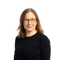 studiokuva ehdokaasta Tiina Davidsainen, hymyilevä nainen, jolla on punertavat hiukset, punertavat silmälasit ja musta pusero 