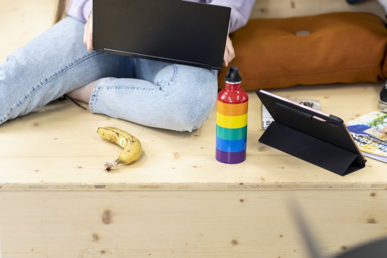 Henkilö istuu portaalla läppäri sylissään, vieressä sateenkaaren värinen juomapullo.