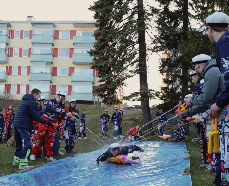 Teekkari laskee maata vasten laitetusta eristemuovista tehtyä liukumäkeä pitkin samalla kun muut ampuvat häntä kohden vesipyssyillä.