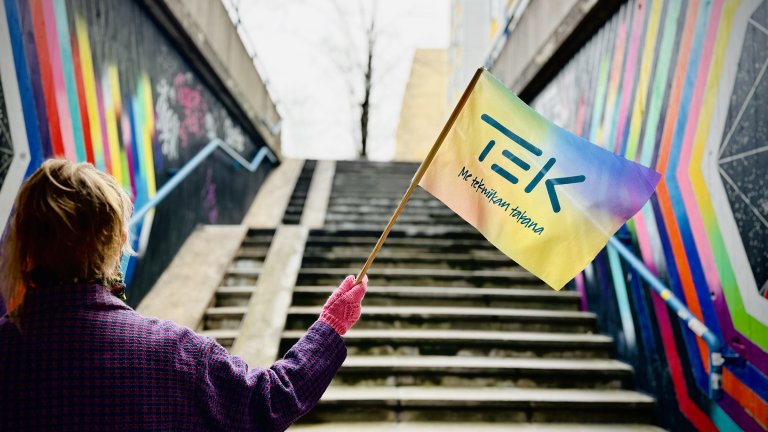 Henkilö heiluttaa TEKin sateenkaarevaa lippua värikkään portaikon alla.