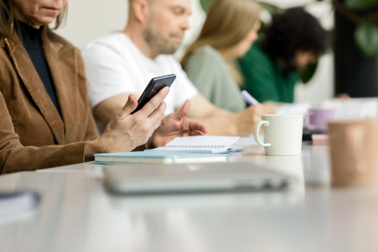 Neljä henkilöä istumassa pöydän ääressä keskittyen omiin tehtäviinsä. Etualalla olevalla on kädessään puhelin ja edessään kannettava tietokone.
