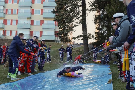 Teekkari laskee maata vasten laitetusta eristemuovista tehtyä liukumäkeä pitkin samalla kun muut ampuvat häntä kohden vesipyssyillä.