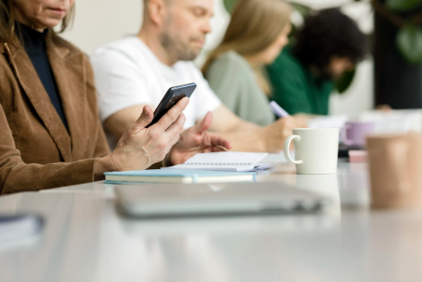 Neljä henkilöä istumassa pöydän ääressä keskittyen omiin tehtäviinsä. Etualalla olevalla on kädessään puhelin ja edessään kannettava tietokone.